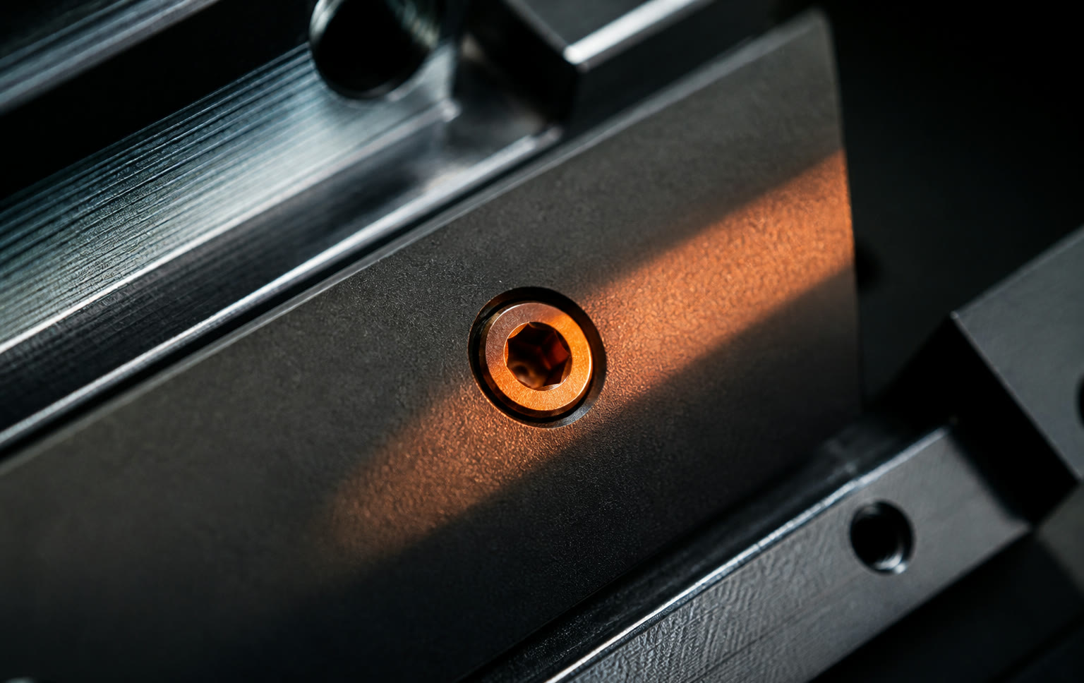 Dark anodized metal plate with one orange hex screw in raking light.